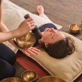 Young woman at peace during sound therapy session. Therapist uses singing bowls for relaxation. Warm lighting on wooden floor enhances tranquil atmosphere