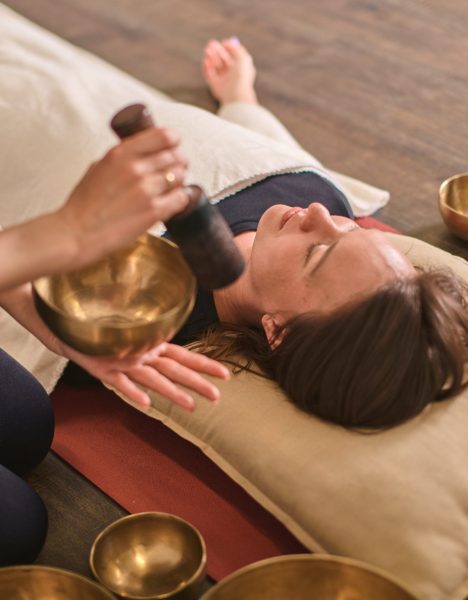 Young woman at peace during sound therapy session. Therapist uses singing bowls for relaxation. Warm lighting on wooden floor enhances tranquil atmosphere