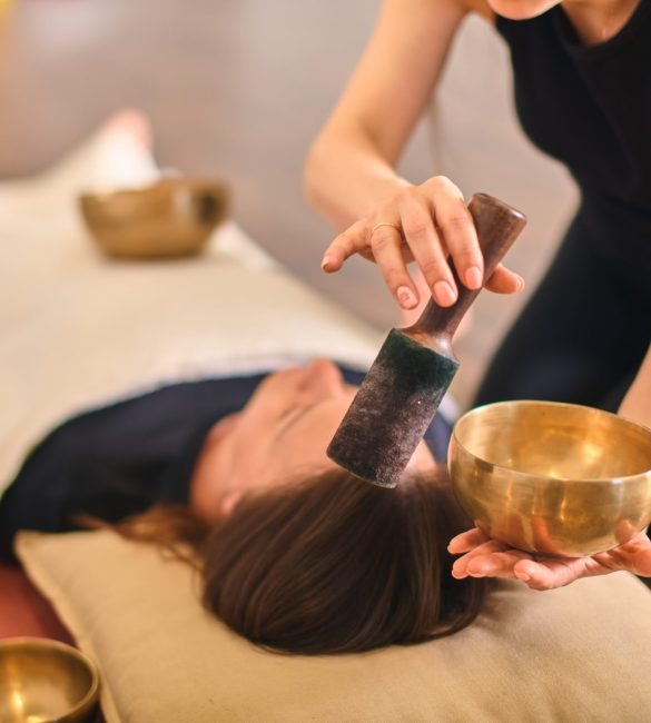 Woman using singing bowl for sound therapy over lying person. Warm lighting enhances calming atmosphere. Bowls placed strategically around for holistic relaxation