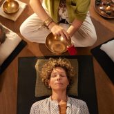 Top view of sound healing session at indoor wellness retreat. Holistic therapist using singing tibetan bell while group lying on back in deep relaxation. High angle view of peaceful moment of energy work, surrounded by crystals and calming atmosphere for people to meditate.